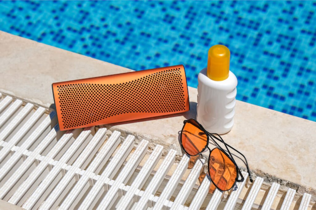 Caixa de som bluetooth perto de uma piscina com um protetor solar e um óculos de sol ao lado